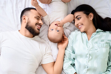 Fototapeta premium Happy young family with their cute little baby lying on bed in bedroom, top view