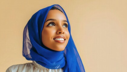 Young woman wearing a bright blue head covering smiles with an upward gaze against a plain background
