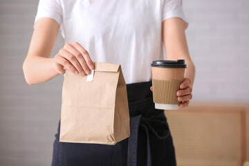 Young female barista with paper bag and cup of coffee in cafe, closeup