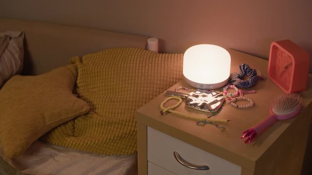 Medium shot of kids single bed with velvet yellow cushions and night stand with lamp, pink clock and scrunchies on top