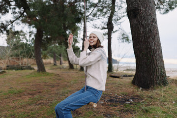 Obraz premium Young girl on a swing beside tall trees in a coastal forest, wearing a knit sweater and beanie, capturing a playful moment in nature.