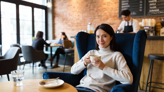 Smiling woman in beige sweater enjoys coffee sitting in comfortable blue armchair at cozy modern cafe with brick wall background. Concept of relaxation, leisure, and urban lifestyle.