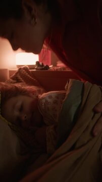 Vertical close up shot of little girl sleeping peacefully while her mother kissing her cheek and adjusting blanket