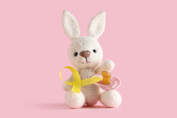Obraz premium Toy bunny, pacifier and yellow ribbon on pink background. Childhood cancer awareness concept