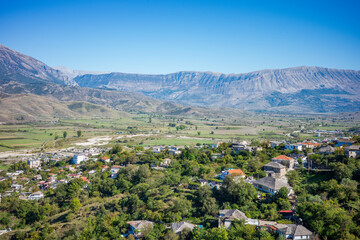 Obraz premium Wide scenic view of a peaceful hillside village overlooking a lush agricultural valley framed by dramatic mountain ranges and a bright blue sky. Traditional houses blend with nature