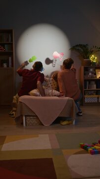 Vertical back view shot of unrecognizable family of three sitting on bed and enjoying shadow puppets