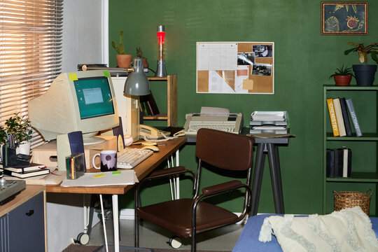 Vintage home office workspace featuring retro computer monitor, keyboard, mouse, telephone, printer and various office supplies arranged on wooden desk against green wall with bulletin board