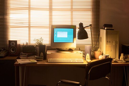 Retro desktop computer setup featuring CRT monitor, tower case, keyboard, mouse, speakers, desk lamp, books, telephone, and potted plant arranged on wooden desk in home office