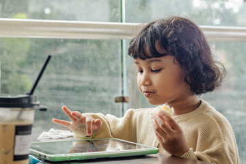 A little Indonesian girl enjoying her breakfast while playing games on her tablet
