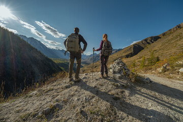 Obraz premium A male and female hiker stand on a high, rocky viewpoint under a bright sun and blue sky, looking out at a vast mountain range and a green valley during an autumn or summer trek.