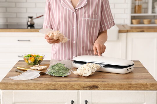 Young woman with vacuum sealer machine for food, parsley and mushrooms in packs on kitchen table