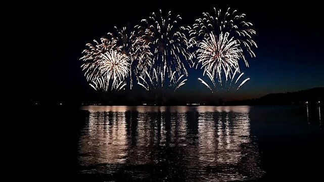Brilliant white fireworks illuminate the dark evening sky above calm reflective water creating spectacular light bursts during a festive night celebration over the horizon.
