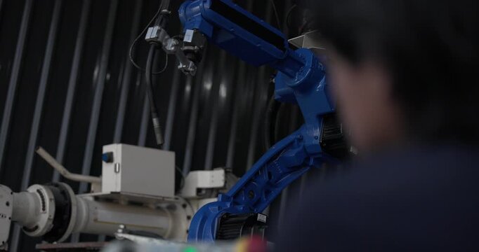 A close-up of a blue industrial robotic arm operating in a factory environment, surrounded by other automation equipment and control panels, highlighting advanced manufacturing technology.
