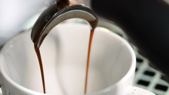 Close up video of fresh espresso coffee flowing from a portafilter into a white cup, professional coffee machine in cafe environment, barista workflow concept, hot beverage preparation.