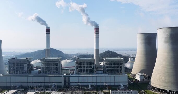 Aerial drone footage panning left across a modern coal-fired power plant: cooling towers, smokestacks emitting steam, and industrial buildings under clear blue sky.