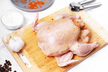 Raw whole chicken seasoned with spices on a wooden cutting board. Prepared poultry with garlic, salt, and pepper ready for roasting on a white kitchen table.