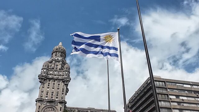 Wehende Nationalflagge von Uruguay vor dem historischen Palacio Salvo in Montevideo bei blauem Himmel mit Wolkenbewegung