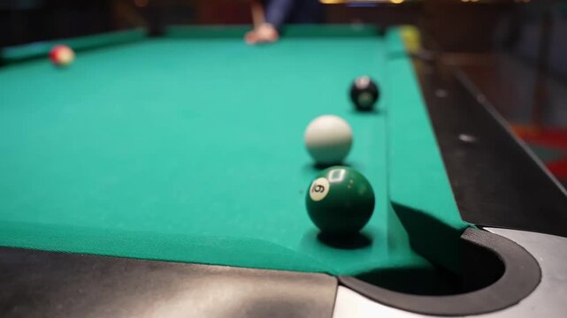 Close-up of a green billiard ball rolling into a corner pocket on a pool table, with cue ball and other balls on green felt, filmed indoors with shallow depth of field.