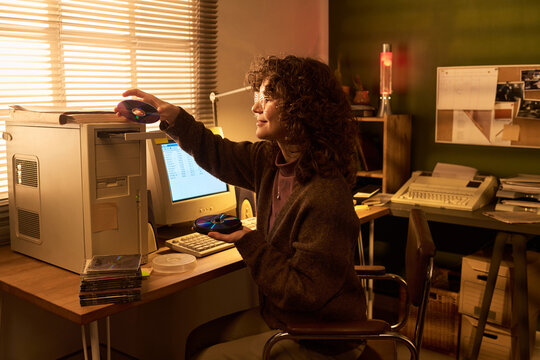 Caucasian woman sitting at desk using floppy disks with vintage computer equipment, holding disk in one hand and inserting another, surrounded by retro office technology