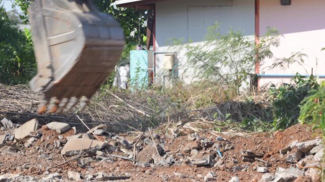 Excavator bucket digging soil, demolition rubble, construction debris cleanup at residential site, clearing broken concrete, brick, metal scrap, dusty earthmoving machinery after renovation