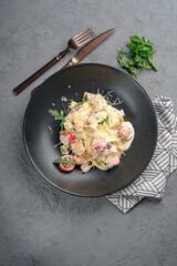 Tagliatelle in creamy sauce with smoked salmon chunks, halved cherry tomatoes, grated cheese and herb sprigs on matte black plate. Indulgent salmon pasta, flavorful gourmet meal