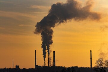 Dark smoke billows from tall industrial smokestacks against a vibrant golden sunset sky, silhouetting the factory skyline and trees below