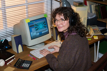 Portrait of Caucasian woman with curly hair wearing glasses and headphones sitting at desk using vintage computer smiling at camera in retro office setting