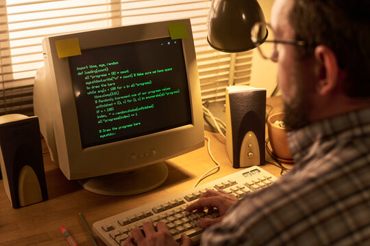 Caucasian middle aged man typing on vintage computer with green text on CRT monitor, sitting at wooden desk with speakers and blinds in background, focusing on programming code