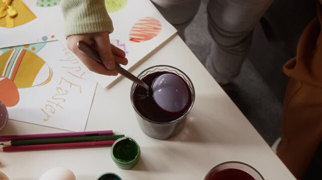 High angle cropped shot of hand of unrecognizable child taking out dyed egg from glass filled with liquid food coloring, preparing for Easter celebration at home