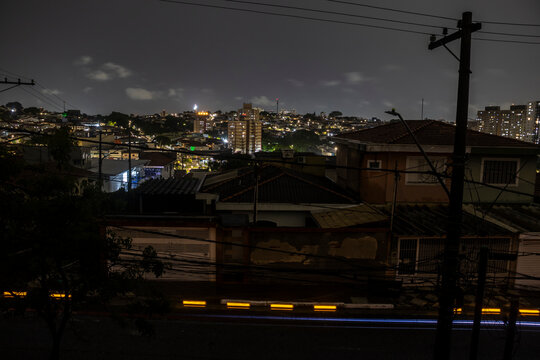 A street in Sao Paulo south side darkens during a blackout. Vulnerable overhead power lines contrast with brightly lit skyscrapers, highlighting the city's deep social and infrastructural divide.