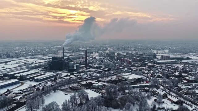 Aerial parallax shot reveals Jelgava urban skyline at sunset featuring central huge industrial plant chimneys emitting plumes amid snow-covered factories buildings trees in Latvia