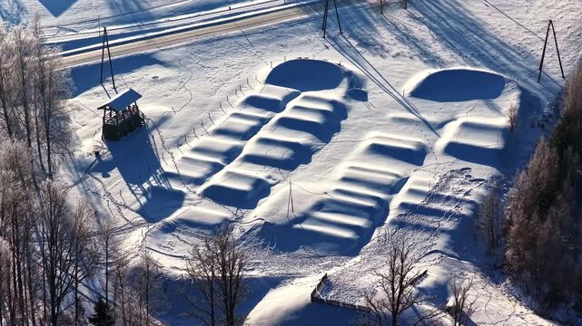 Aerial parallax of a snow-blanketed dirt jump bike park in Vecpiebalga, Latvia, featuring white berms and jumps under sunrise light with frost-covered birch trees and long shadows.