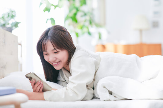 日差しが入るベッドルームでスマホを見るパジャマを着た若い女性