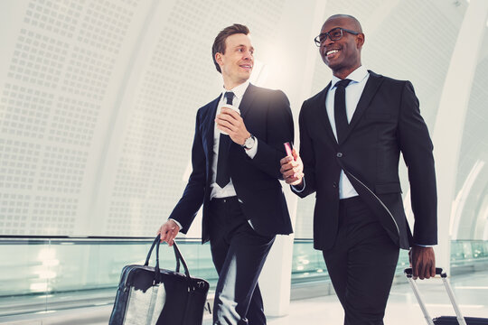 Smiling diverse executives walking down an airport departure hall talking together. They are traveling on a business engagement, with a cabin-sized suitcase and passport in hand.