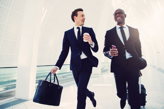 Smiling diverse businessmen running down an airport departure hall. They are traveling on a business engagement, with a cabin-sized suitcase and passport in hand. 
