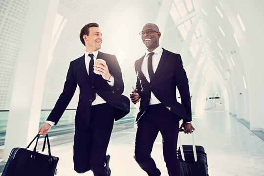 Two smiling diverse businessmen running down an airport departure hall. They are traveling on a business engagement, with a cabin-sized suitcase and passport in hand.