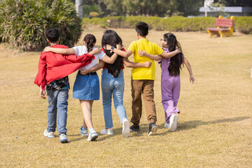 Obraz premium Group of indian children friends walking with arms around each other enjoying time together outdoor in summer park. Asian kids having fun. Childhood. Back view.