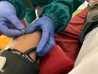 Medical professional wearing blue gloves inserting needle into patient. Person lying on medical chair with intravenous drip attached to their arm for blood transfusion or donation.