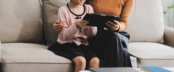 A cheerful Asian mother helps her young daughter do homework and color a book at home, while the father works on a laptop in the background. A cozy, bright, and modern family lifestyle.