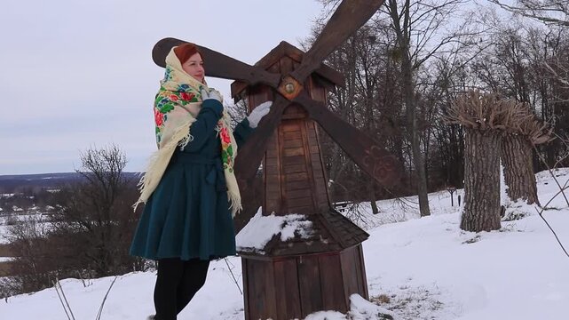 Beautiful redhead woman in winter coat and bright scarf standing near wooden mill on winter background