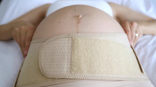 Expectant mother lying in bed adjusting a maternity support belt on her big belly to help with back pain. Pregnancy wellness. Orthopedic care. And prenatal support for a comfortable gestation period