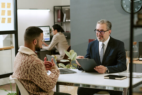 Middle aged Caucasian man wearing glasses interviewing young adult man in modern office, both sitting at desk with laptop and clipboard, colleague working in background