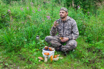 A European man is drinking tea in the forest. There is a bucket of berries and mushrooms nearby.