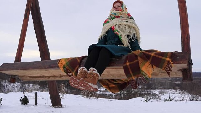 Beautiful girl swinging on a large swing with a view of a winter village and snowy fields