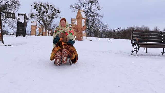 Beautiful girl sledding on a snowy slope among wooden buildings. Winter outdoor recreation, active day and atmospheric snowy landscape.