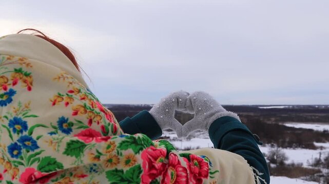 Girl in a scarf shows a heart with her hands against the background of a winter landscape