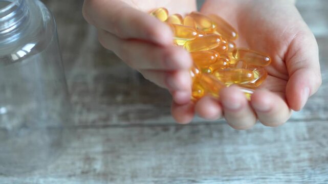 Hands holding soft gel capsules above an empty glass jar on a wooden table, showcasing the process of transferring supplements in a bright indoor setting