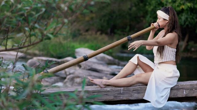 A serene moment by the water as a woman skillfully plays a bamboo Instrument didgeridoo, showcasing both grace and musicianship in a natural outdoor setting.