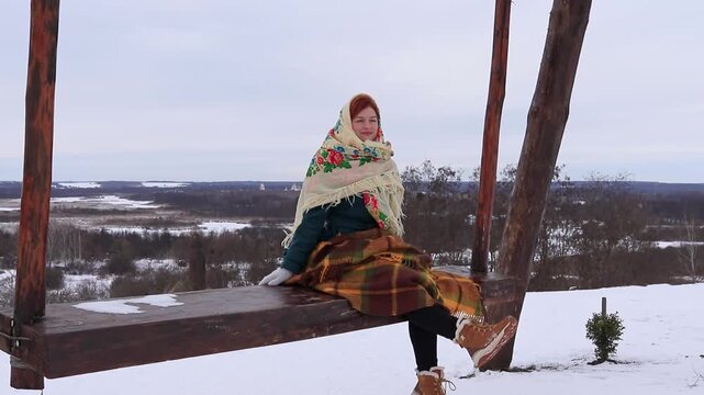 A woman sits on a wooden swing amidst a snowy landscape. A peaceful winter landscape, cold air and atmospheric mood in nature.