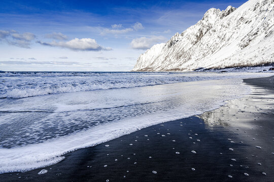 View of the dark, glistening beach reflecting the crisp, foamy waves as they meet the snow-dusted mountains under a bright, clear sky, Ersfjord Senja, Nordland, Norway.